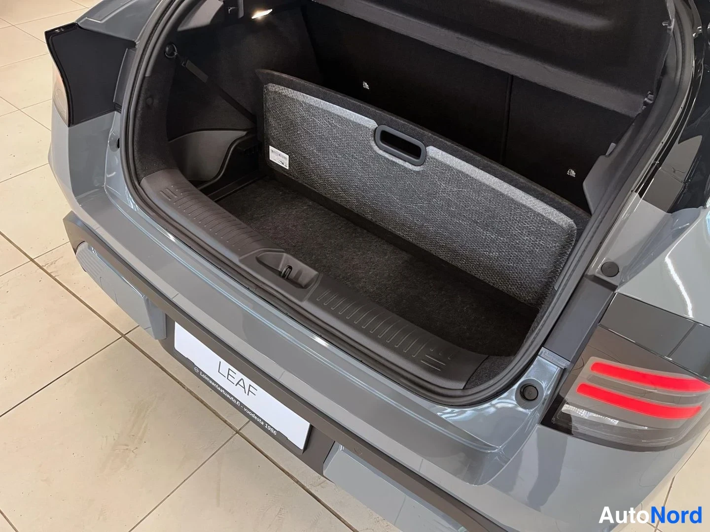 Tavaratila: Close-up of the cargo area floor in a grey Nissan Leaf showing a removable carpeted floor mat or panel.