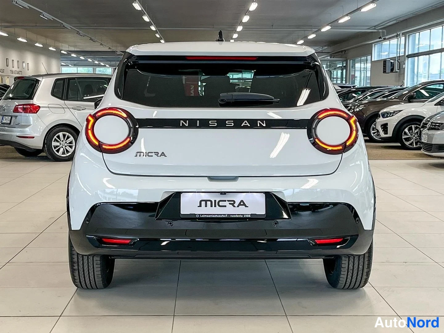 Takaa: Valkoinen Nissan Micran takapää suoraan takaa valokuvattuna autosalissa; pyöreät takavalot, musta korokela jossa NISSAN- ja MICRA-tekstit.