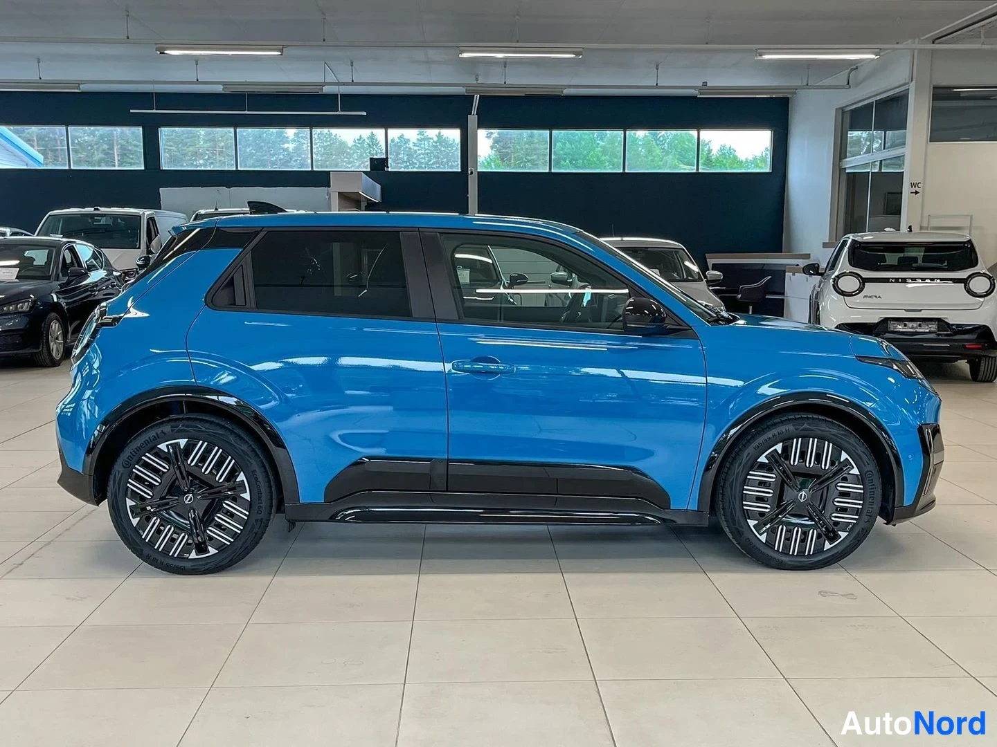 Sivulta: Sininen Nissan Micra sivulta vasemmalta, näkyy koko auto, musta katkaisukiinnike ovenkahvoissa ja tummat alumiinivanteet.