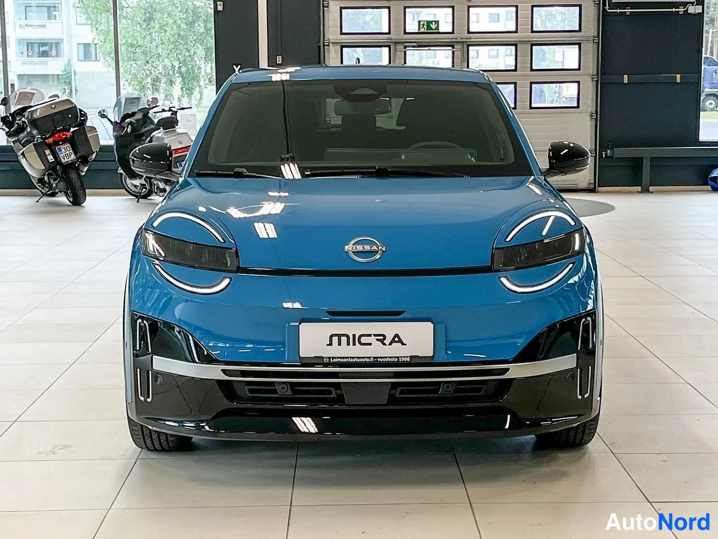 Edestä: Sininen Nissan Micra suoraan edestäpäin, näkyvät Nissan-merkki, ajovalot, MICRA-tunnus etusäleikössä ja etupuskuri.