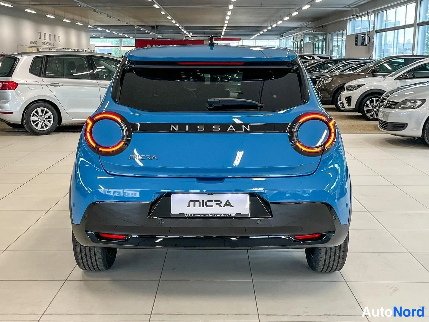 Takaa: Sinisen Nissan Micran takanäkö showroomissa: pyöreät takavalot, MICRA-koriste ja tumma takapuskuri.