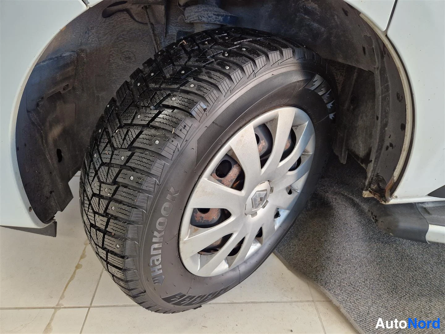 Muu: Lähikuva autossa olevasta nastarenkaasta (Hankook) hopeisella vanteella, näkyy Renault-logo ja likainen pyöränkotelo.