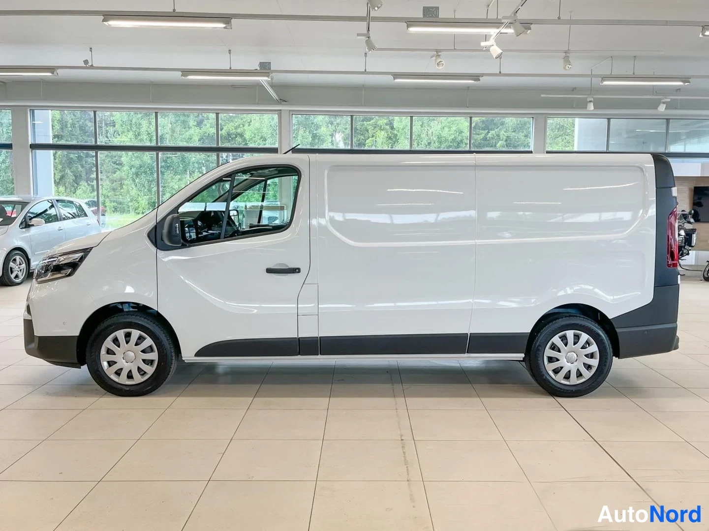 Etuviisto, nokka vasemmalle: Valkoinen Nissan Primastar -pakettiauto sivulta oikealta, liikehallissa.