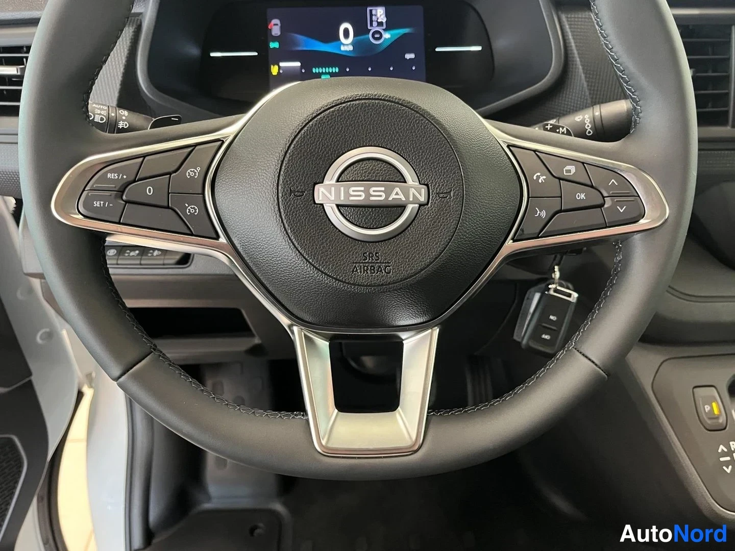 Sivulta: Nissan-merkkinen ohjauspyörä, jossa monitoimanapit ja SRS AIRBAG -teksti; takana osittain digitaalinen kojetaulu.