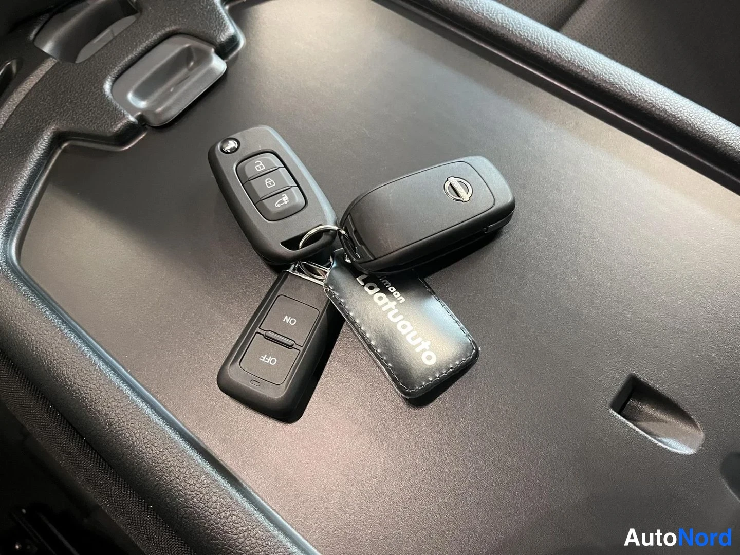 Yksityiskohta: Kaksi ajoneuvon avainnippua ja Laatuauto-avainnappi tummanharmaalla matolla.