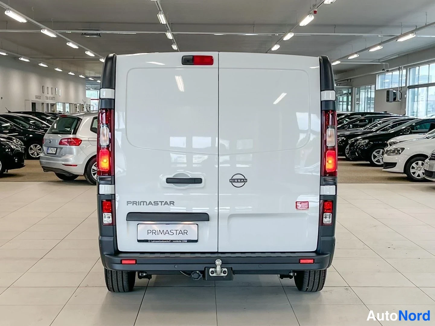 Takaa: Auton takanäkymä: valkoinen Nissan Primastar -pakettiauto, takavalojen osa palaa, takaluukissa PRIMASTAR-teksti ja Nissan-merkki, takassa vetoconfiguring.