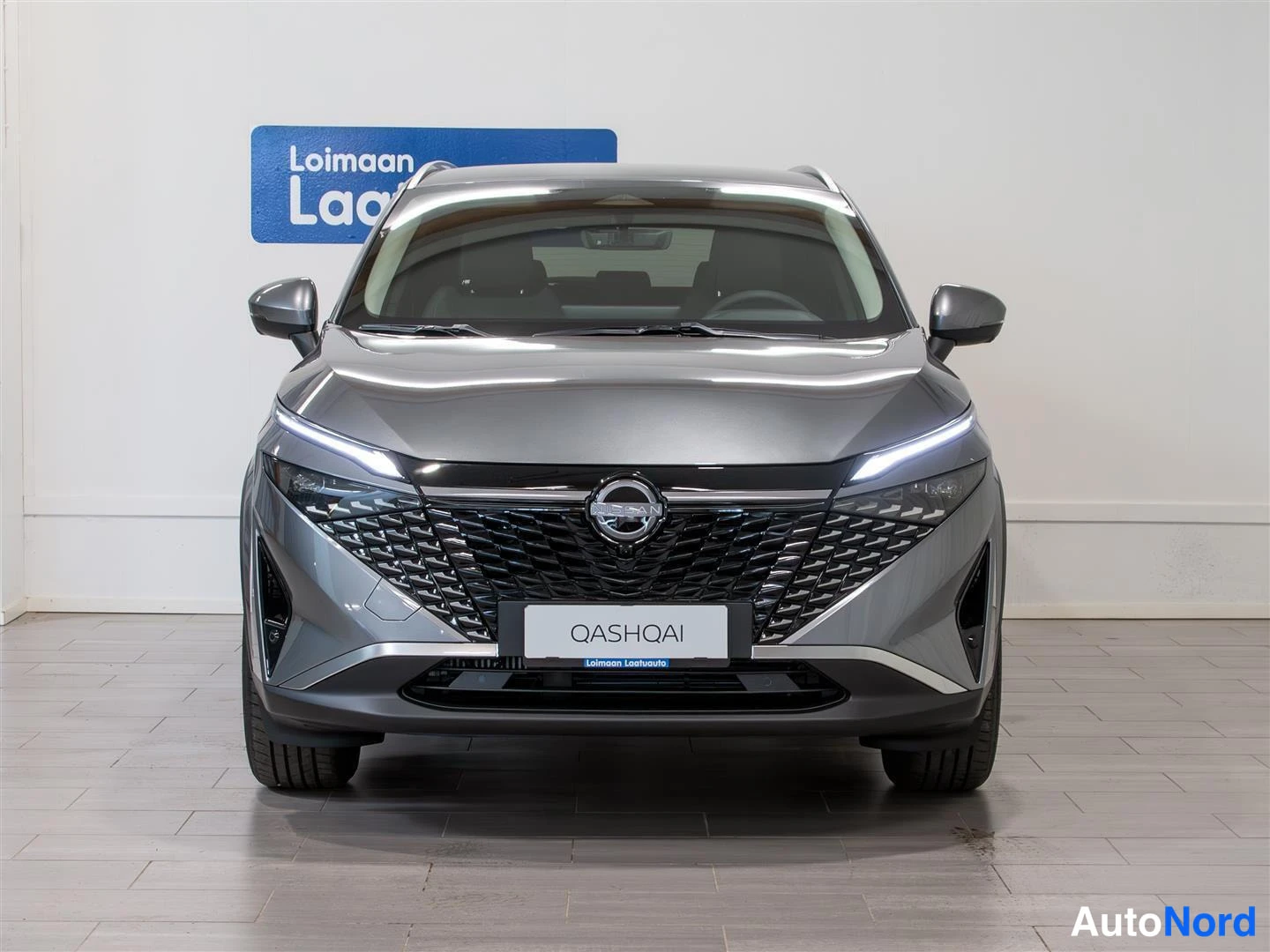 Edestä: Nissan Qashqai -mallin suora etunäkymä keskellä V-muotoista etusäleikköä, ohut LED-etuvalot ja QASHQAI-kyltti etupuskurissa harmaassa showroomissa.