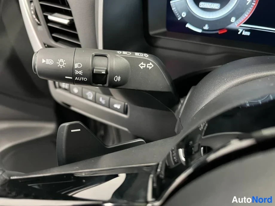Yksityiskohta: Ohjauspyörän valon- ja suuntavilkkujakytkimet AUTO-merkinnällä ja LED-symbolilla; mittariston Standard-näkymä taustalla.