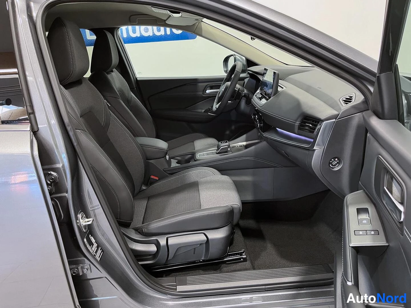 Etuviisto: Nissan Qashqain etumatkustustila: tummat kankaanistuimet, ohjauspyörä ja kosketusnäyttö keskellä kojelautaa, matkustajan ovi auki.