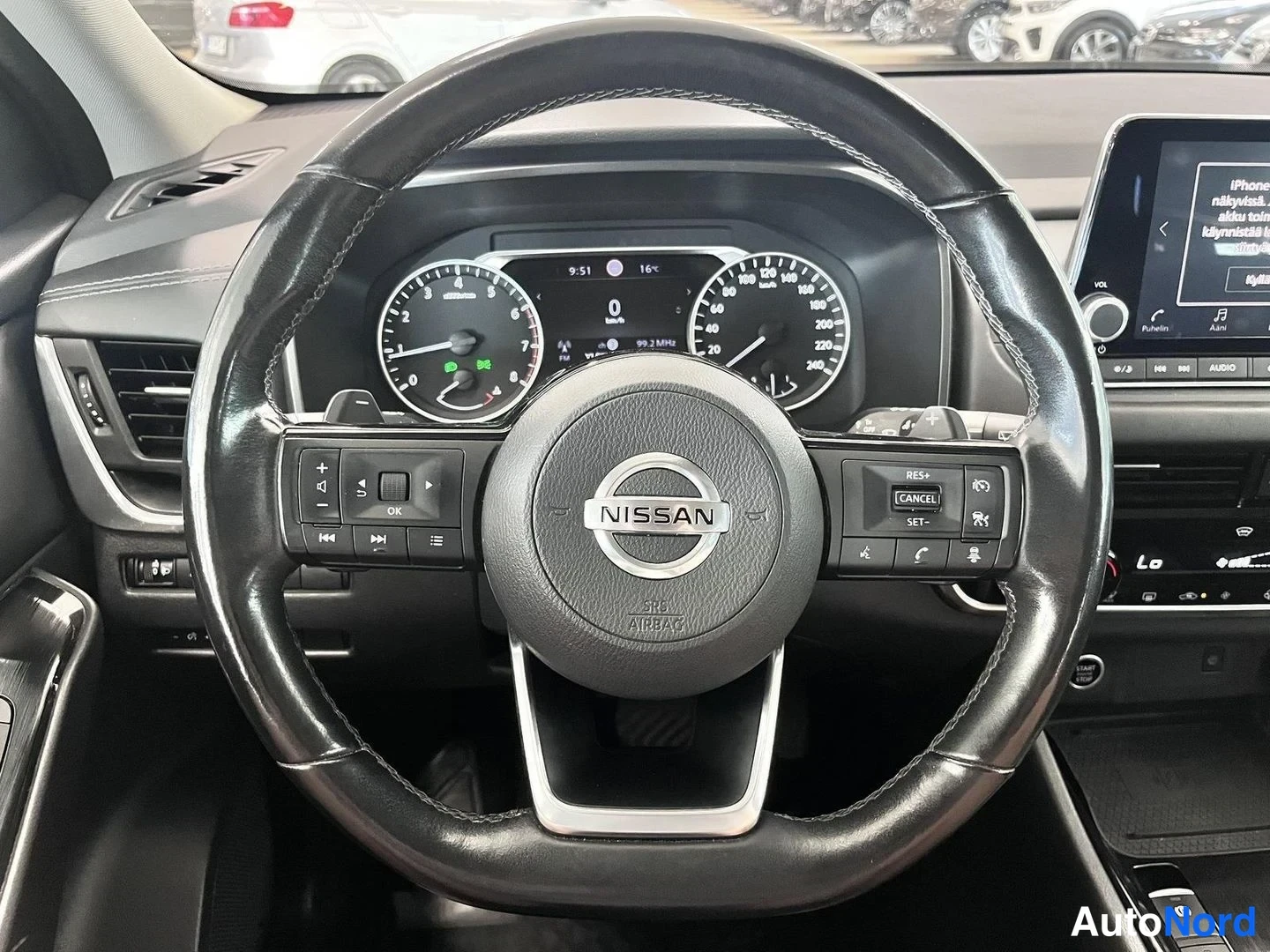 Yksityiskohta: Ohjauspyörä Nissan-merkillä, ympärillä mittaristo ja osa infotainment-näytöstä ja ilmastoinnin merkinnöistä (Lo). Ohjauspyörän napit sisältävät risteilyssäätime