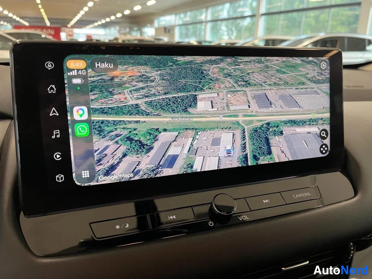 Muu: Auton kojelaudan suuri kosketusnäyttö, jolla näkyy Google Maps -karttasovellus ilmasateliittikuvassa; näytön reunassa sovelluskuvakkeita ja alla äänensäätimet s