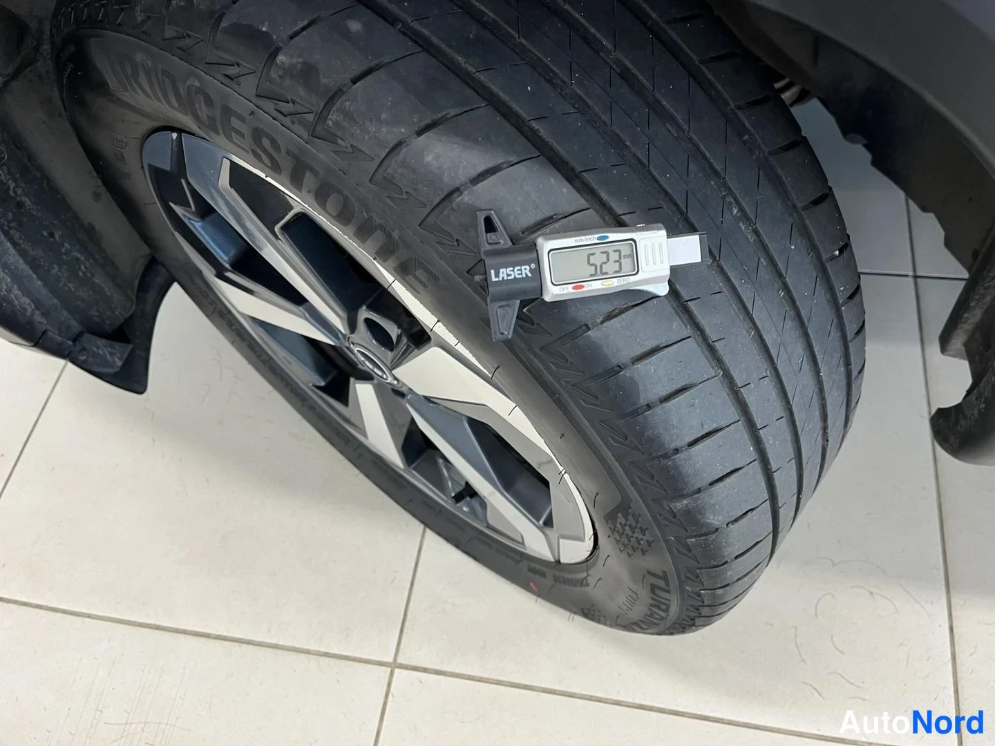 Yksityiskohta: Auton rengas Laser-tuotemerkin pyöränpölymittarilla, näyttö lukemassa 5,23 mm; Bridgestone Turanza -rengas ja harmaa alumiinivanne valkoisella laatoitetulla lat
