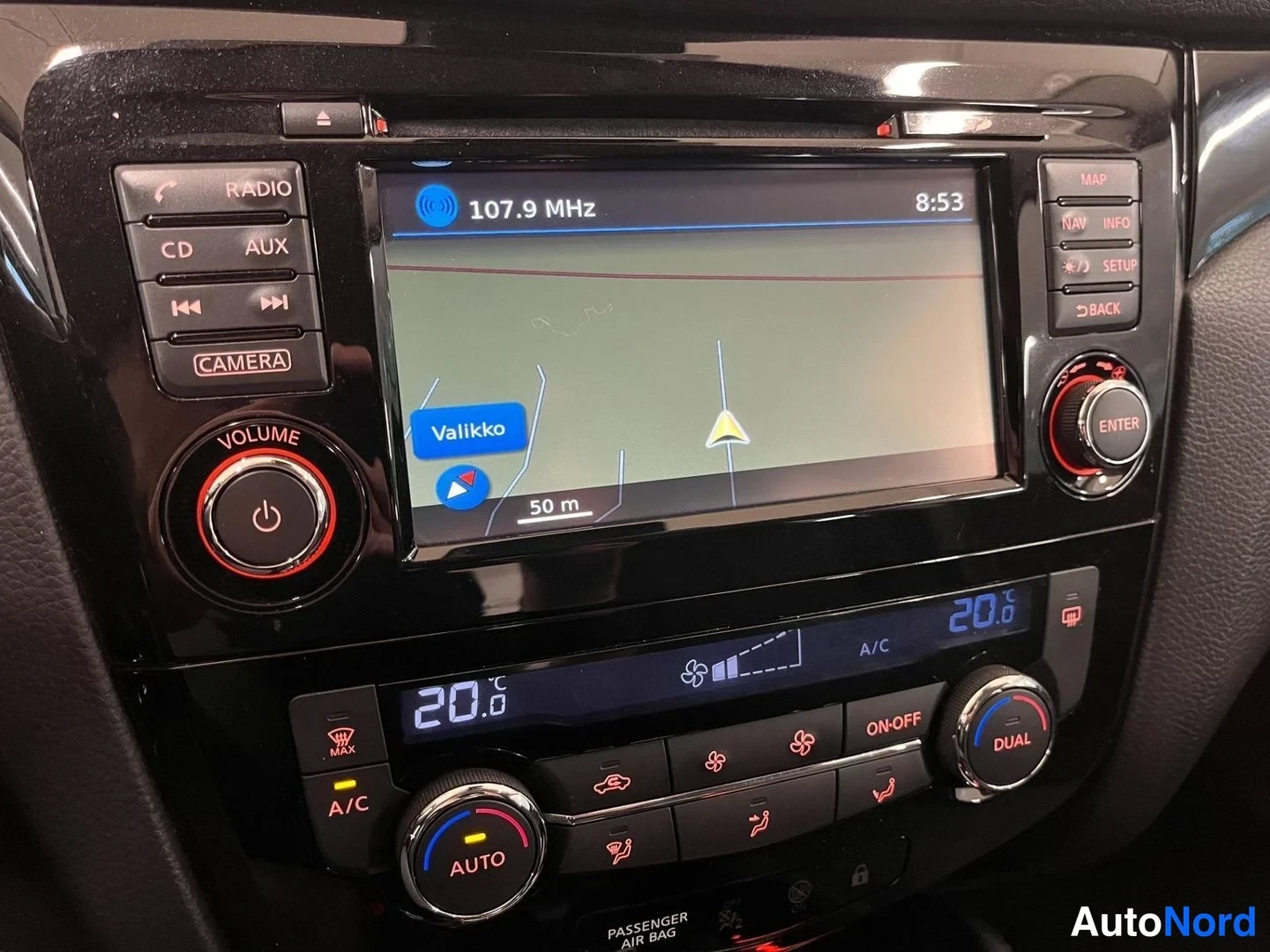 Yksityiskohta: Radiopainikkeet, kamera-nappi ja volume-kiekko vasemmalla, kartta radiofrekvenssillä 107.9 MHz ja aika 8:53 ylhäällä; ilmastointi- ja navigointipainikkeet ylhää