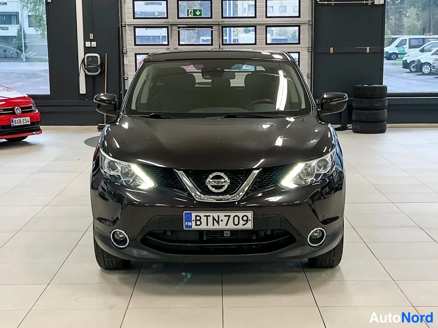 Edestä: Tummanvioletti Nissan Qashqai -henkilöauto suoraan edestä, päiväajovalot päällä, rekisterikilpi.