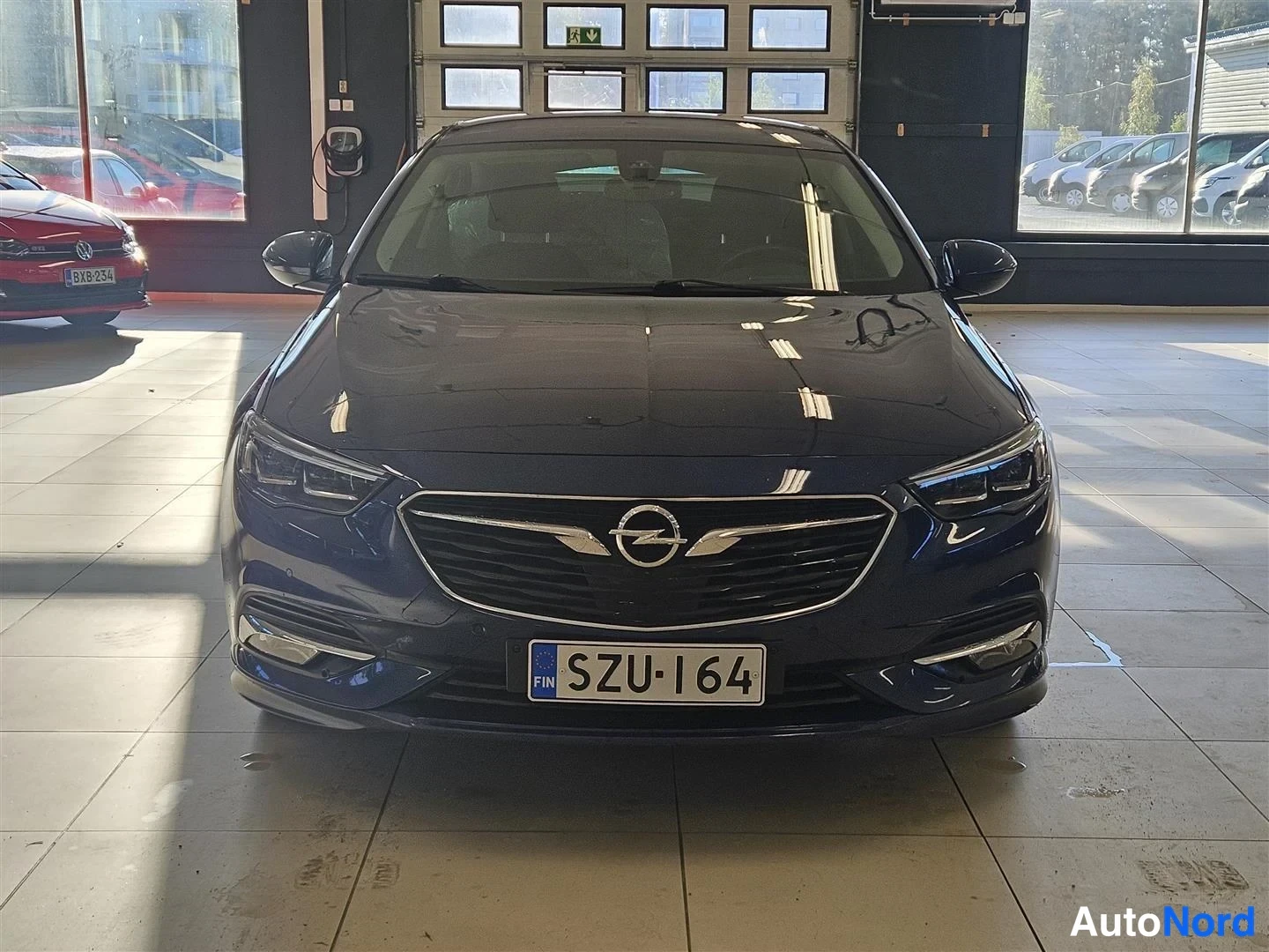 Edestä: Tummansinisen sedanin suora etukuva näyttämötilassa; Opel-merkki, LED-ajovalot ja rekisteritunnus näkyvät.