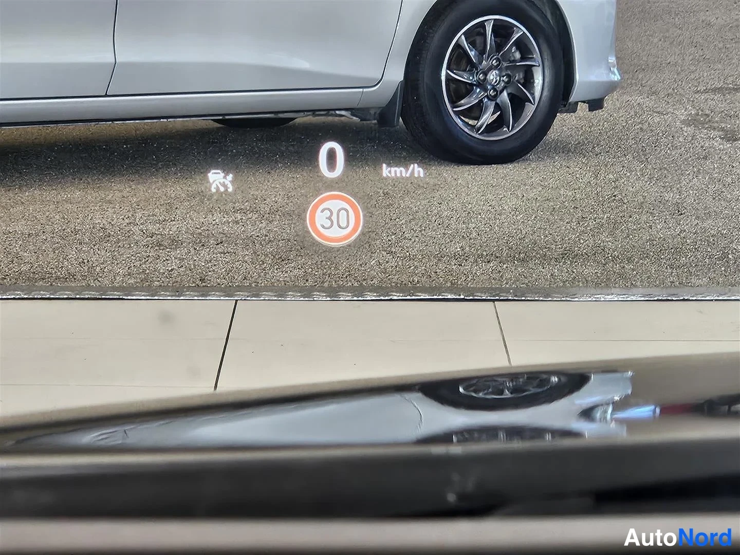 Yksityiskohta: Tuulilasiin heijastuva head-up -näyttö, jossa lukema 0 km/h ja liikemerkki 30; taustalla hopeisen auton etupyörä ja kori.