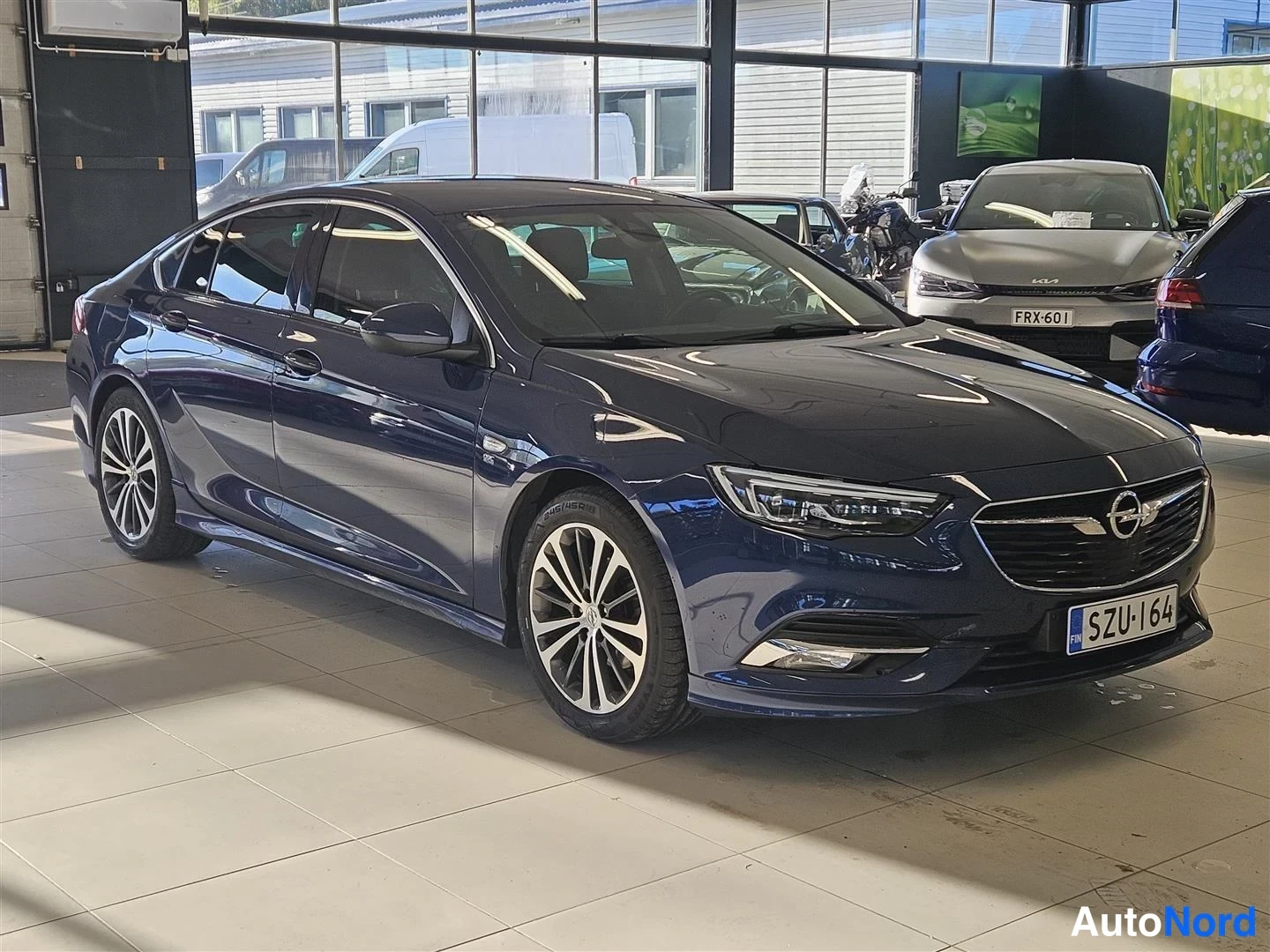 Takaviisto: Tummansinisen sedanin takakulman kuva sisätiloissa; kannen takavalot, Opel-merkki ja rekisteritunnus näkyvät.