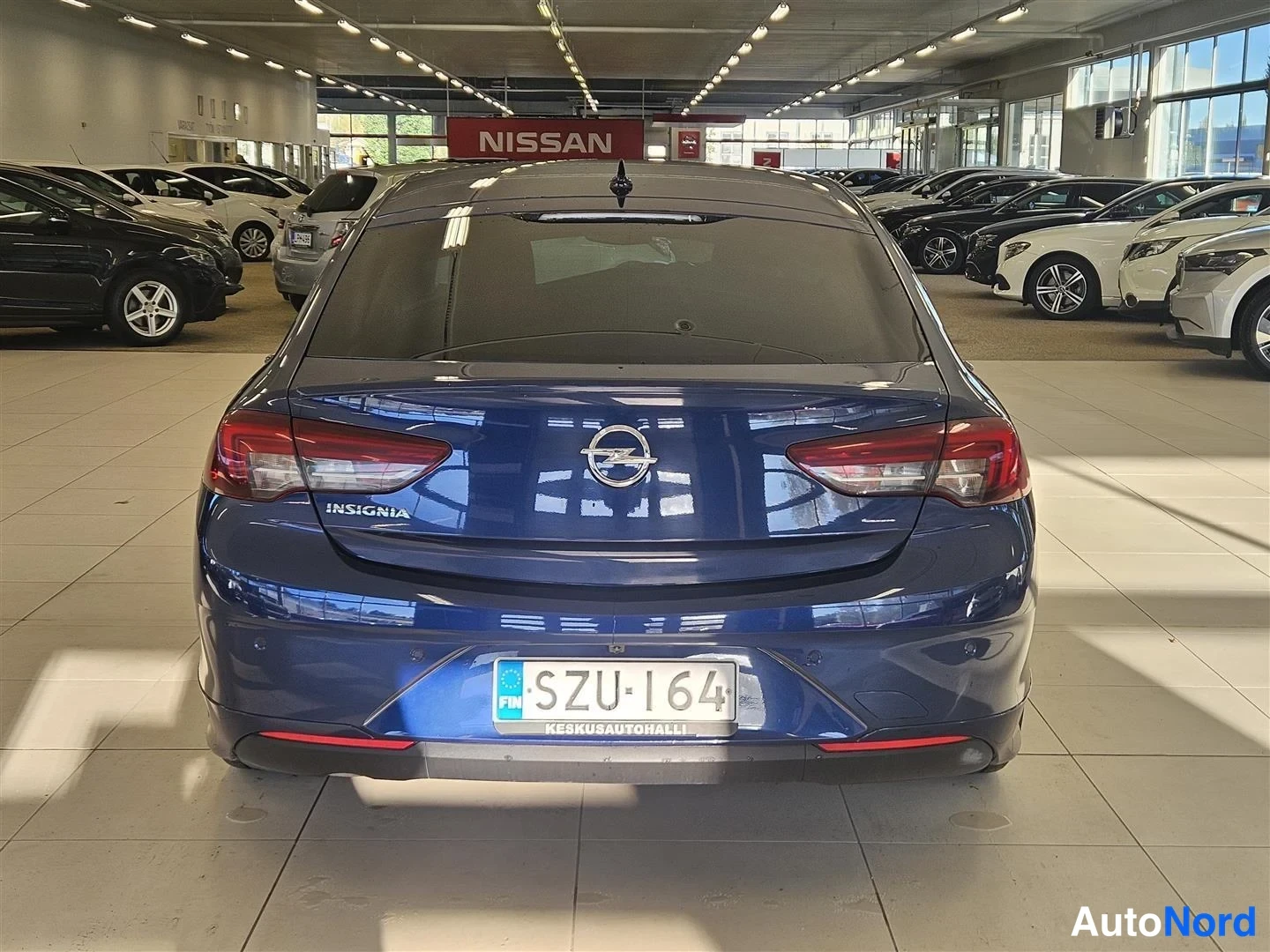 Takaa: Opel Insignian takaosa keskittyneesti; rekisterikilpi ja Keskusautohalli-tarra, sininen kori autosalissa.