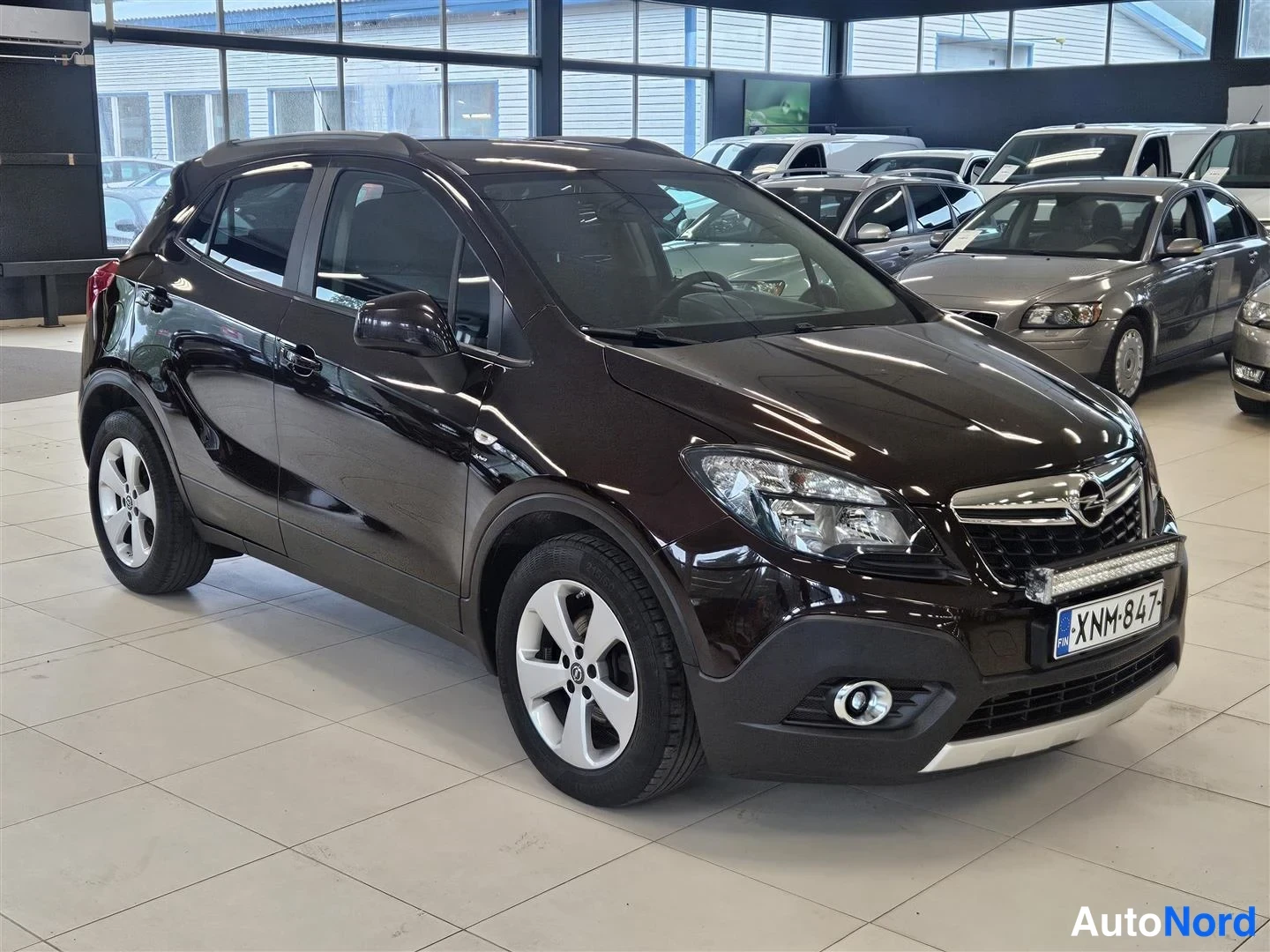 Etuviisto, nokka oikealle: Opel Mokka tummanruskeana etuviistosta oikealta, rekisteri, LED-palkki puskurissa, autosalon lattialla muiden autojen joukossa.