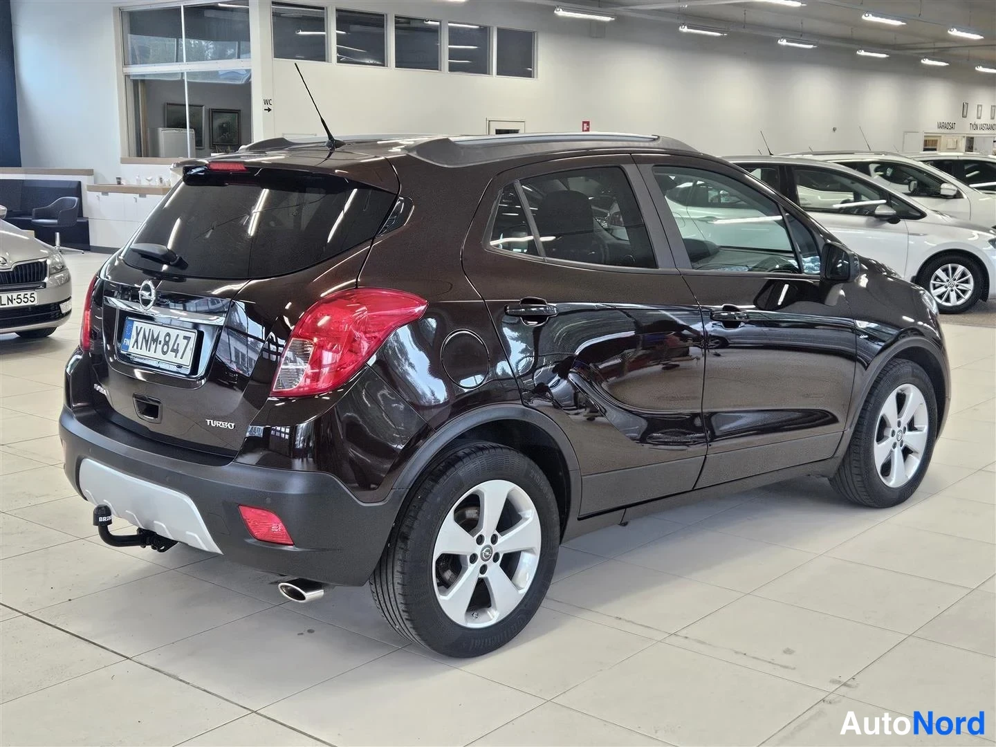 Takaviisto: Opel Mokka takaviistosta oikealta, TURBO-teksti takaluukussa, vetokoukku ja BOSAL-merkki, rekisteri, autosalon sisätiloissa.