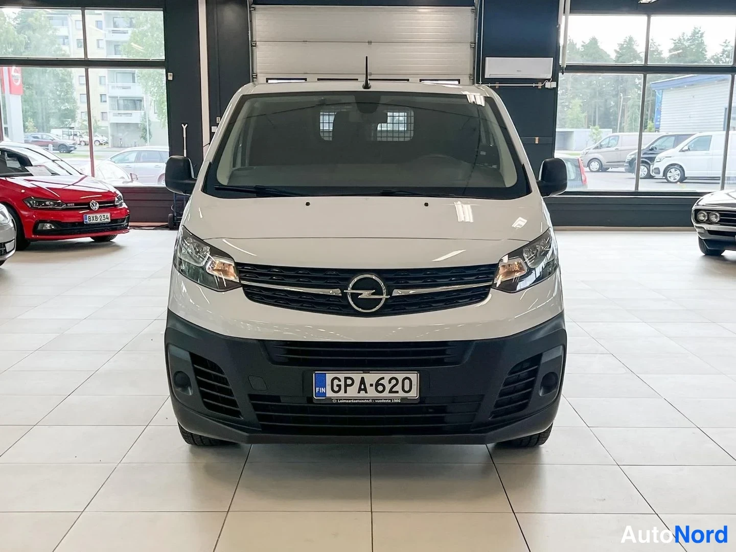 Edestä: Valkoinen pakettiauto suoraan edestä nähtynä autonäyttelytilassa, rekisteritunnus ja Opel-merkki säleikössä.
