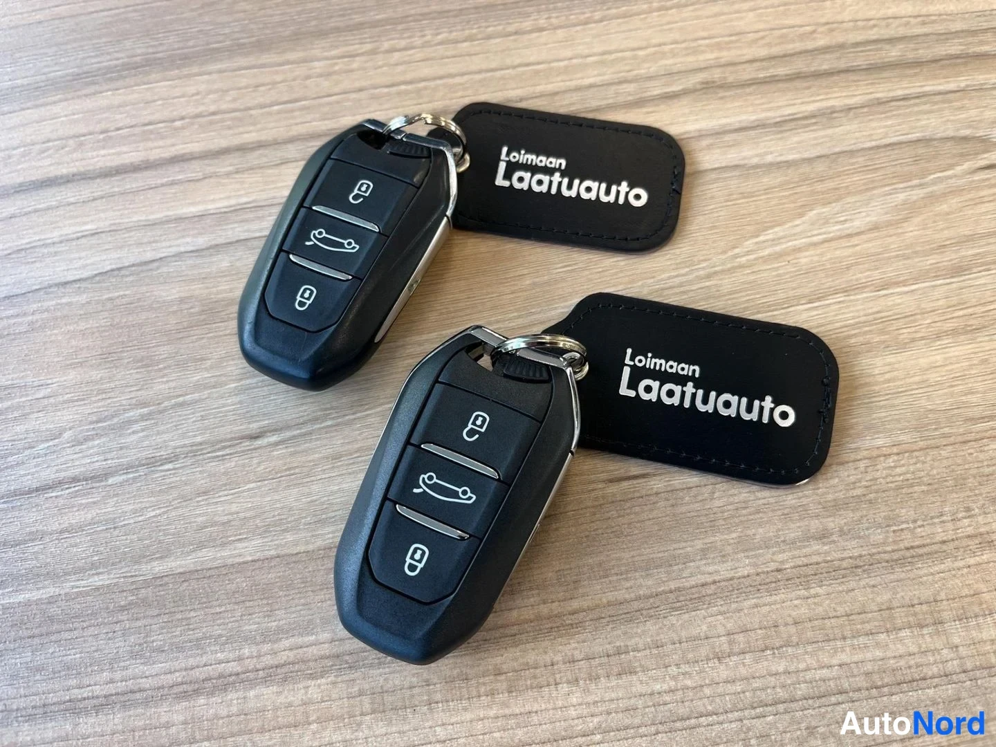 Yksityiskohta: Kaksi Peugeoton kauko-ohjaimia ja mustat avainniput, joissa teksti Loimaan Laatuauto, puupöydällä.