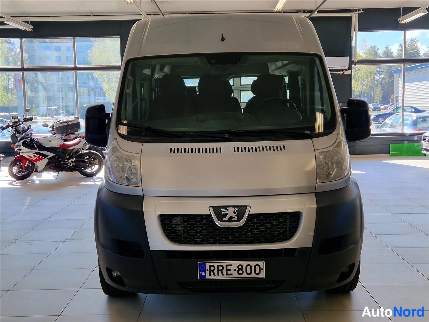 Edestä: Peugeot Boxer -pakkun etupääsuoraan edestä kuvattuna automohteerin sisätiloissa; Peugeoton merkki, etusäleikkö ja rekisterikilpi näkyvät, taustalla moottoripyör