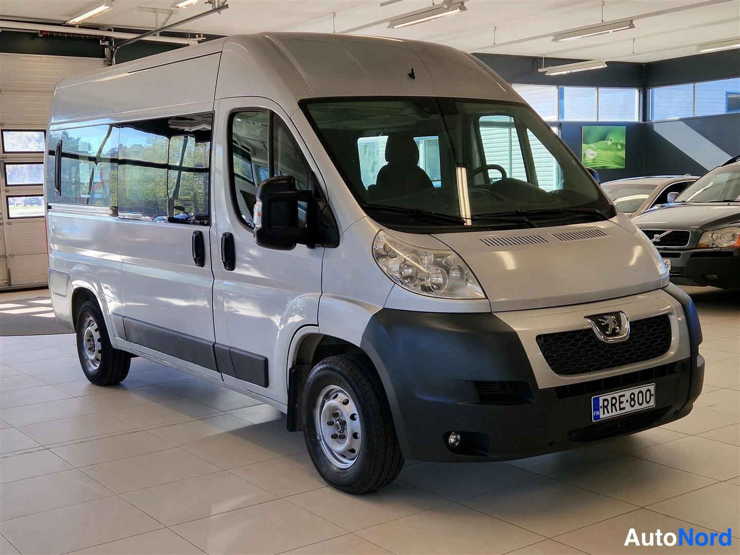 Etuviisto, nokka oikealle: Hopeanharmaa Peugeot Boxer -pakettibussi kolmannelta etu-nurkkakulmalta, nokka oikealla; kyljen ikkunat, Peugeoton merkki ja rekisterikilpi näkyvät autom