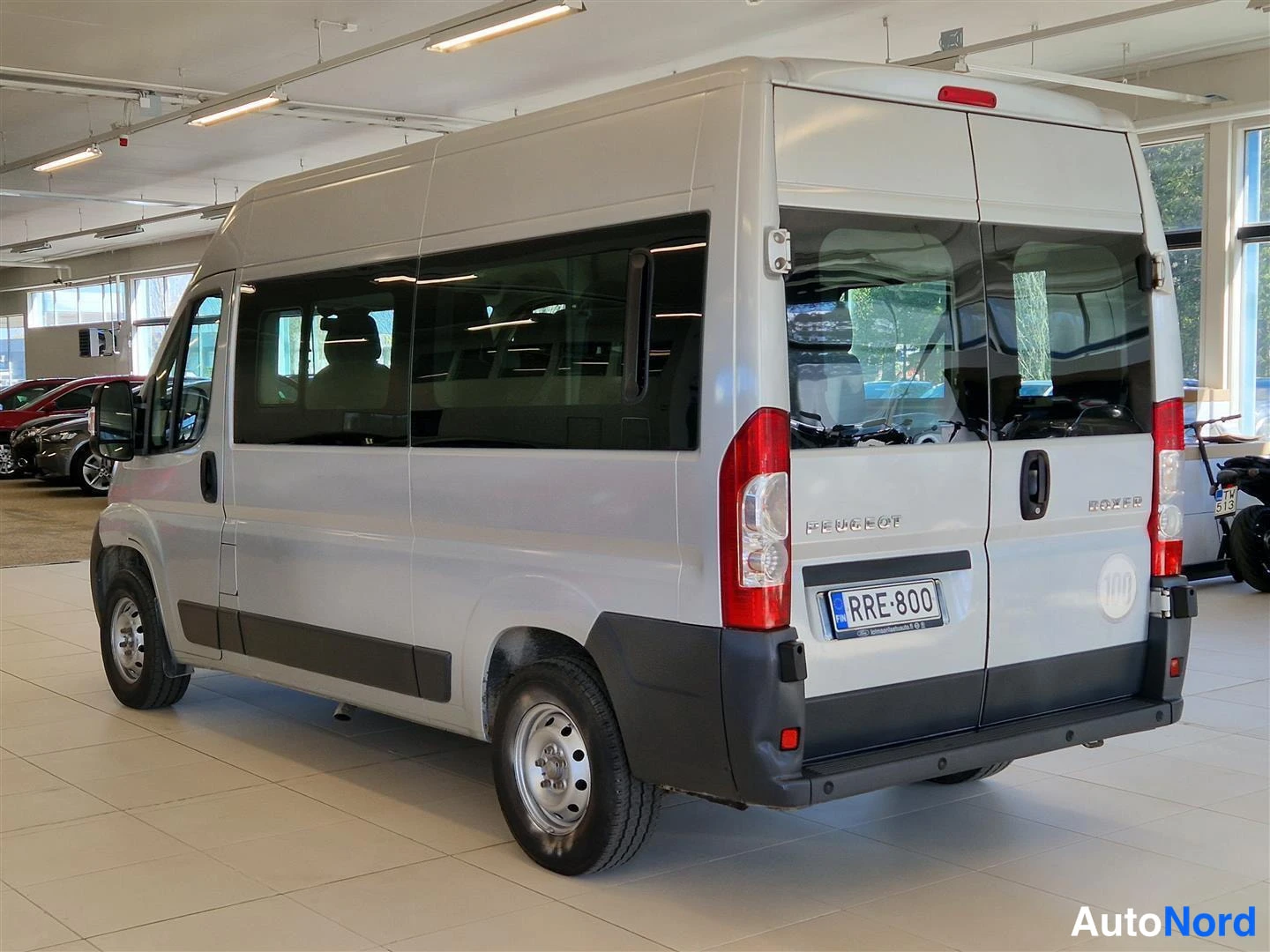 Takaviisto: Peugeot Boxer -pakettiauto vasemmalta takaosalta kuvattuna autonäyttelyhallissa; tummat ikkunat ja rekisterikilpi.