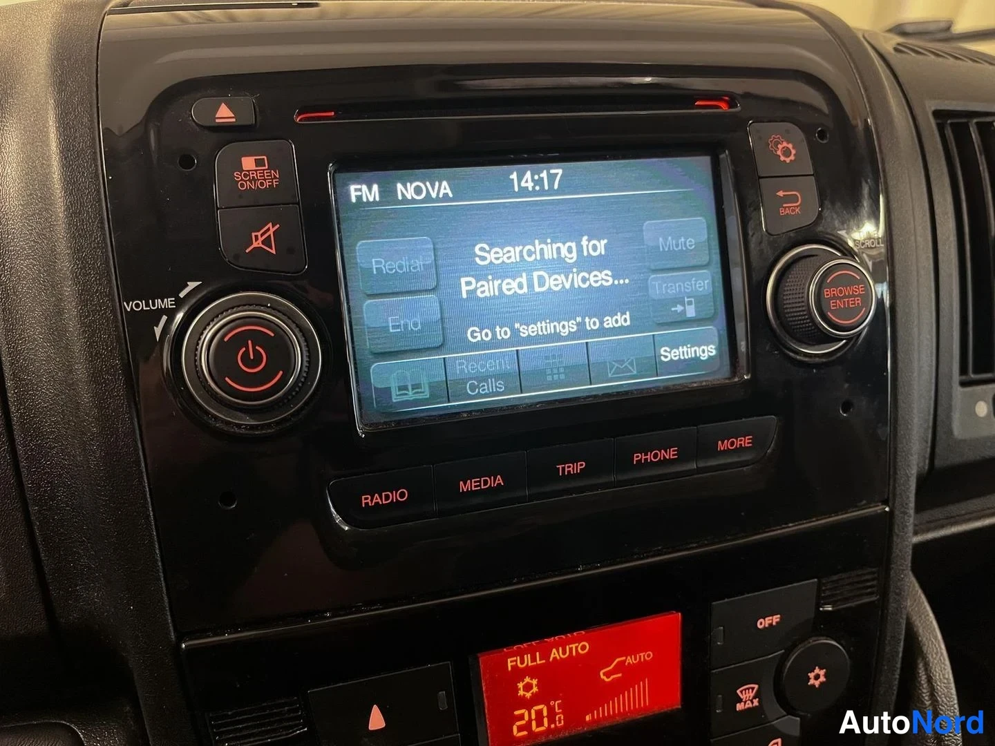 Yksityiskohta: Auton infotainment-näyttö radiokanavalla FM NOVA kello 14:17, ruudulla teksti 'Searching for Paired Devices...'; kellubuttonit RADIO, MEDIA, TRIP, PHONE, MORE j