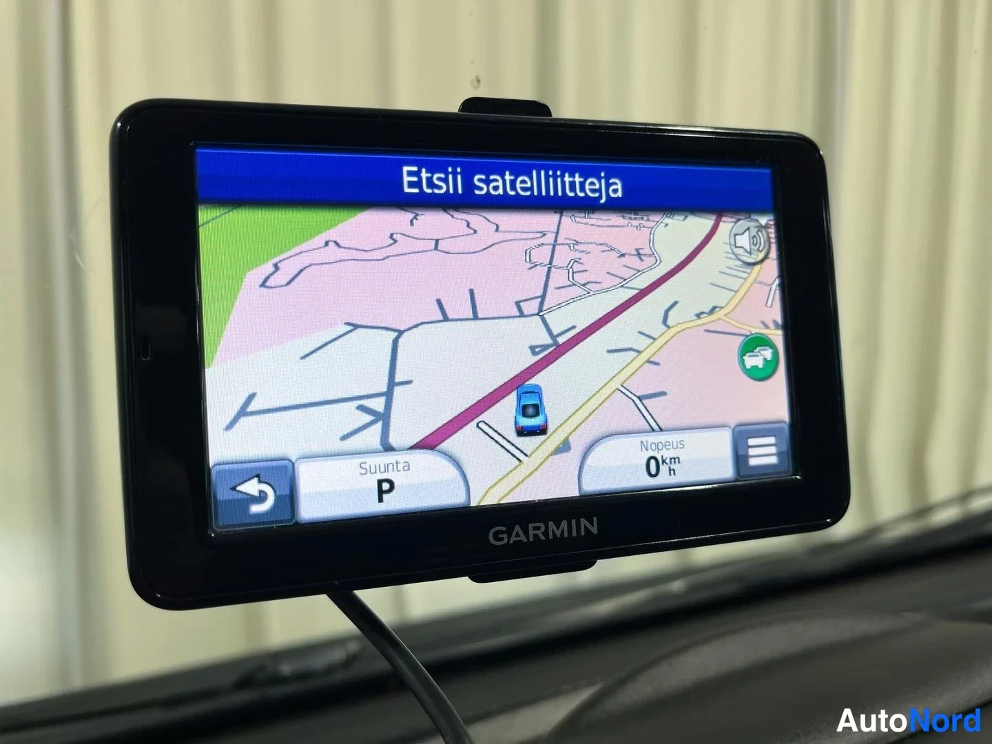 Yksityiskohta: Kiinnitetty Garmin-navigaattori, jonka näytöllä teksti 'Etsii satelliitteja' ja kartta; nopeus 0 km/h.