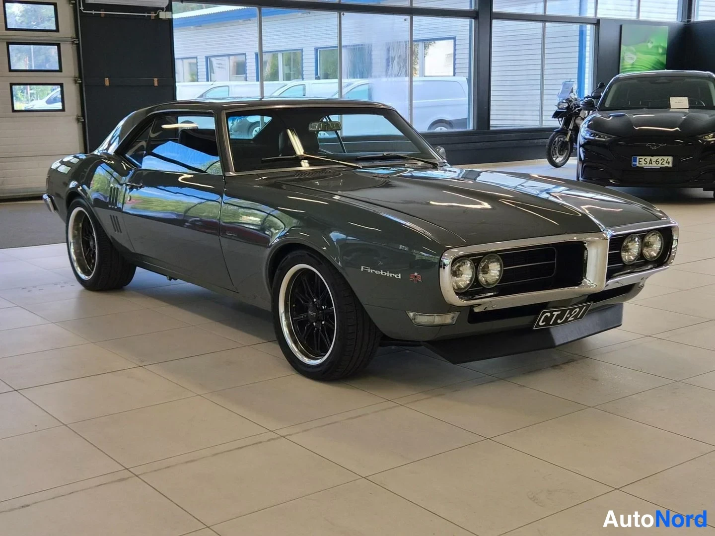 Etuviisto, nokka oikealle: Harmaa Pontiac Firebird -auto etuosa ja vasen sivu näkyvissä autonäyttelytilassa, rekisteritunnus.
