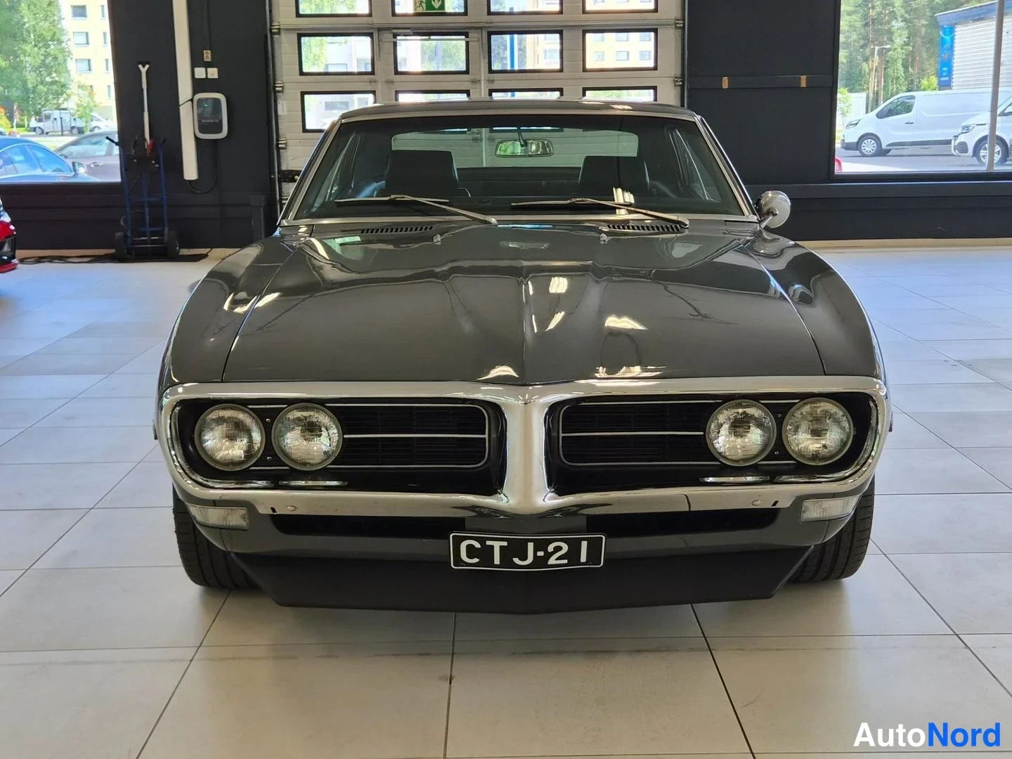 Edestä: Harmaan Pontiac Firebird -auton etunäkökula sisätiloissa, etusäleikön ja neljästä pyöreästä ajovalosta näkyy, rekisteritunnus.