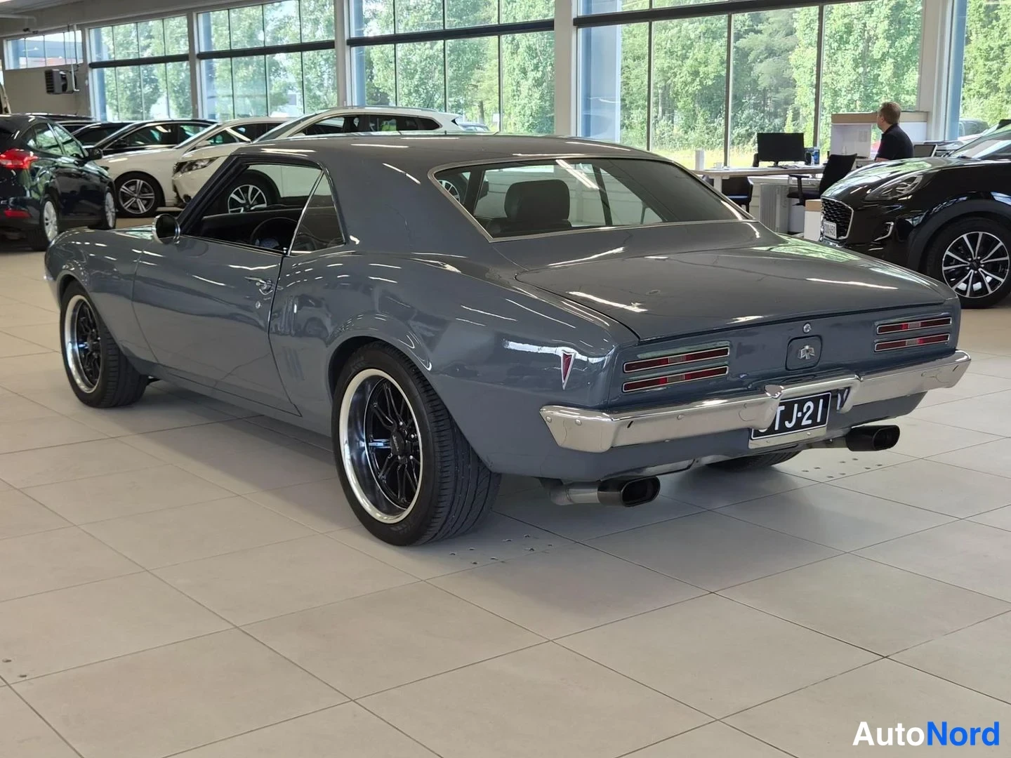 Takaviisto: Harmaansininen Pontiac Firebird -coupe takaviistosta autonäyttelysalissa; takavalot, kromipuskuri ja -rekisterikilpi näkyvissä.