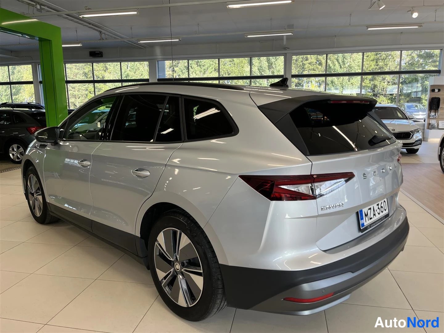 Takaviisto: Skoda Enyaq iV -sähköauto hopeanvärisenä takanurkkauksesta vasemmasta taustasta, rekisterikilpi, autonäyttelysalissa.
