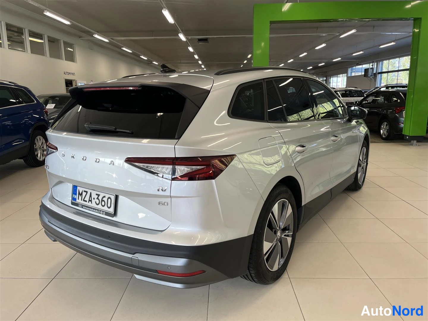 Takaviisto: Skoda Enyaq 60 iV -sähköauto hopeanvärisenä takanurkkauksesta vasemmasta taustasta, rekisterikilpi, autonäyttelysalissa.