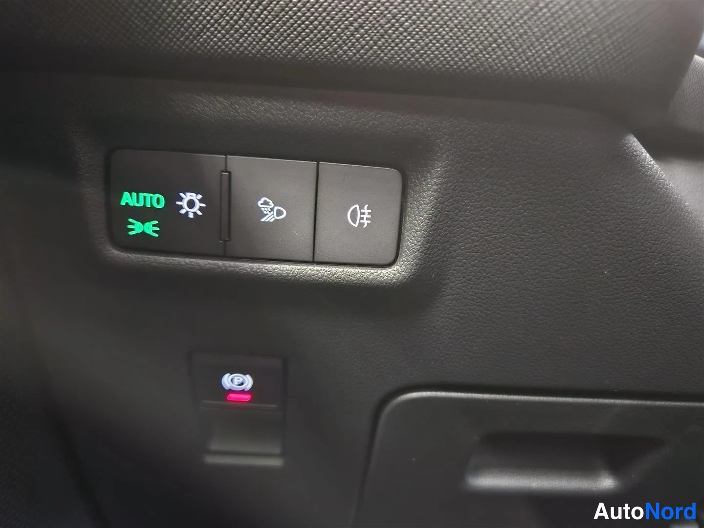 Yksityiskohta: Autojen sisätilan valokytkinpaneeli, jossa AUTO-teksti ja sumuvalojen merkki ovat vihreinä, sekä alla punainen valo.