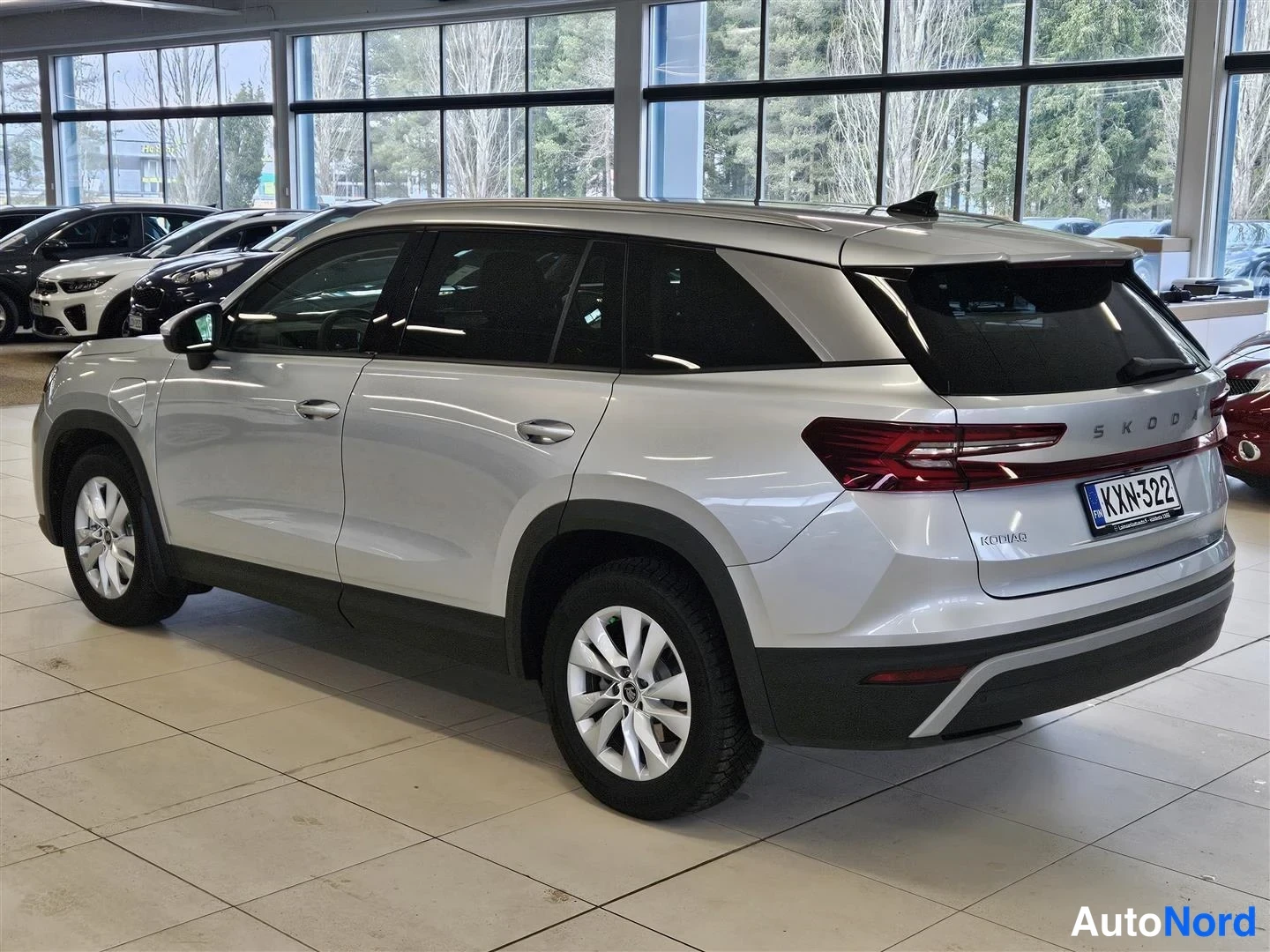 Takaviisto: Hopeanvärinen Škoda Kodiaq -maasturin takakulmanäkymä lasiseinän edessä, takaosaan SKODA- ja KODIAQ-tekstit, rekisterikilpi.
