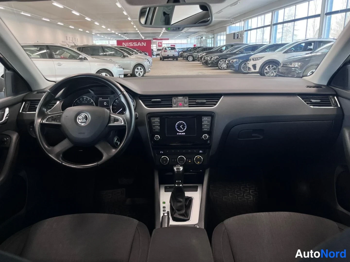Muu: Ohjaamo etusytytty: Skodan ohjauspyörä, infonäyttö, automaattivaihteen valitsin ja tummat penkit; showroom näkyy takalasin läpi.