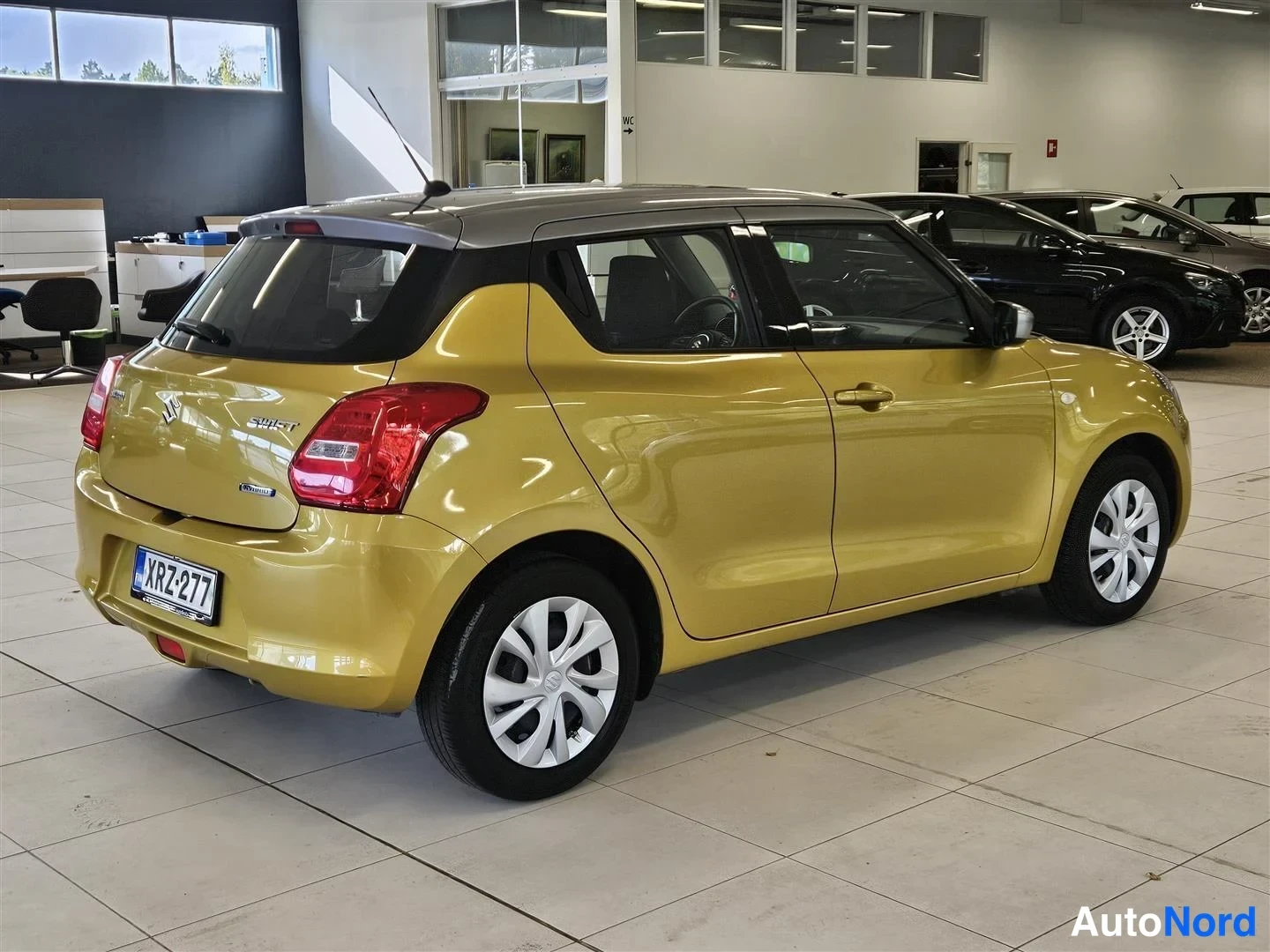 Takaviisto: Keltainen Suzuki Swift viistosti takaa leftalta nähtynä; rekisteritunnus, tumma katto ja takavalojen punainen muotoilu näkyvät myymälätilassa.