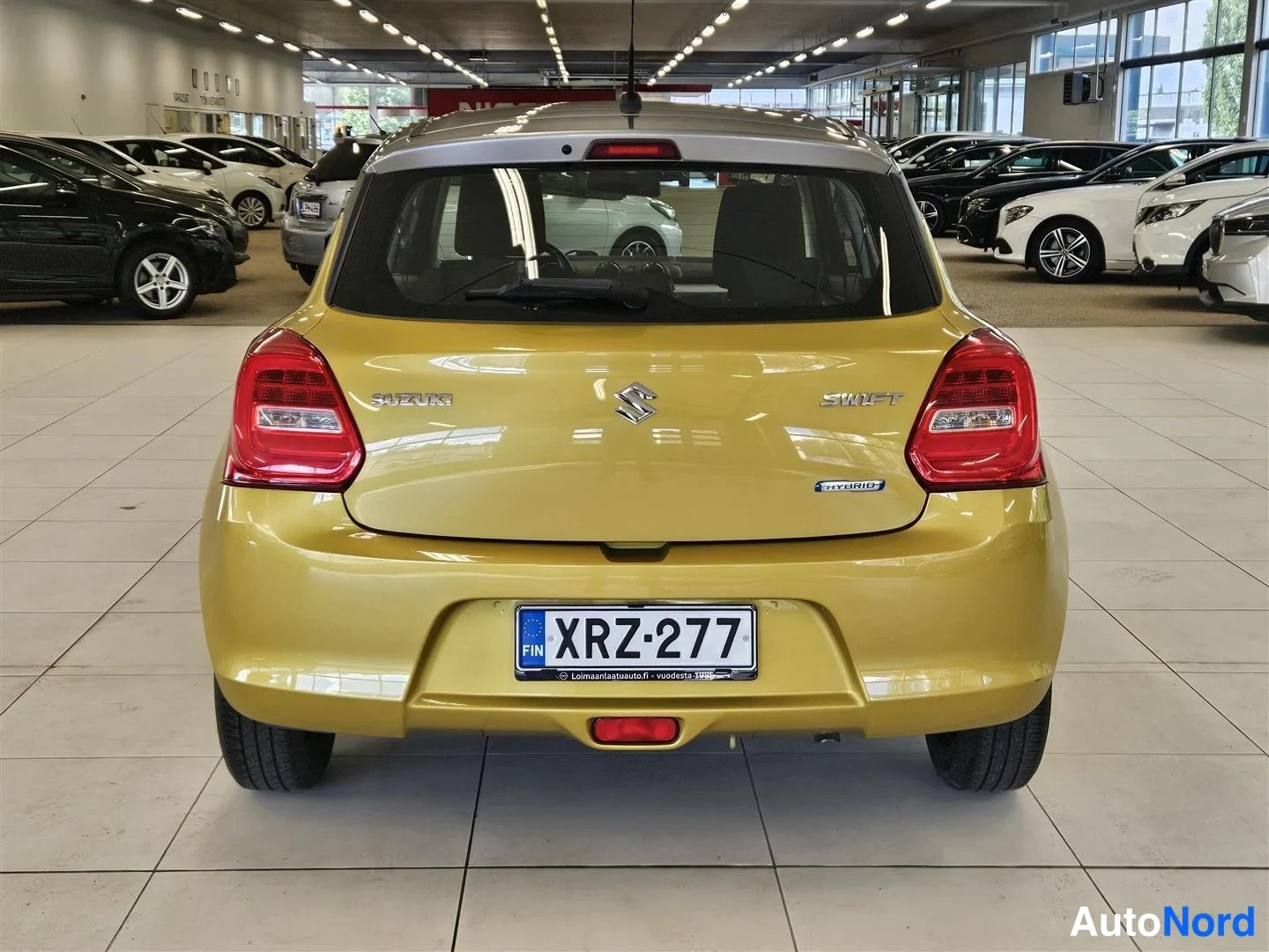 Takaa: Keltaisen Suzuki Swiftin takaosa suoraan takaa nähtynä; tavaratilan kannessa Suzuki- ja Swift-merkit sekä Hybrid-tarra, rekisteritunnus.