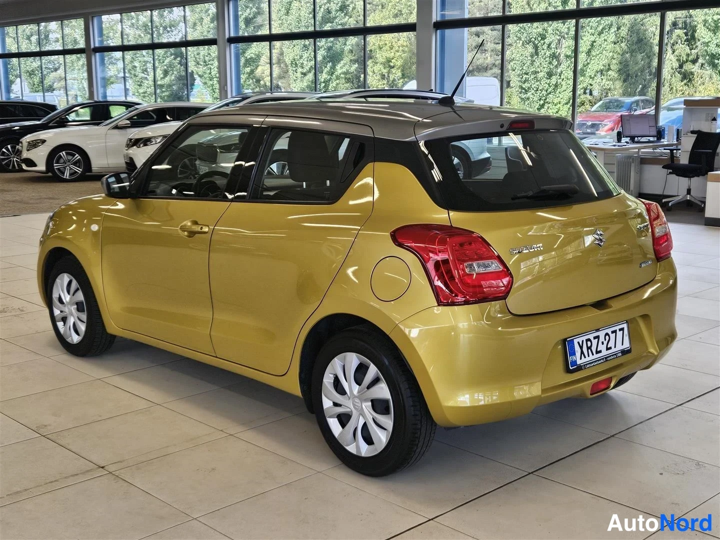 Takaviisto: Keltainen Suzuki Swift viistosti takaa oikealta nähtynä; rekisteritunnus, tumma katto ja Swift-merkki tavaratilan kannessa näkyvät autohallissa.