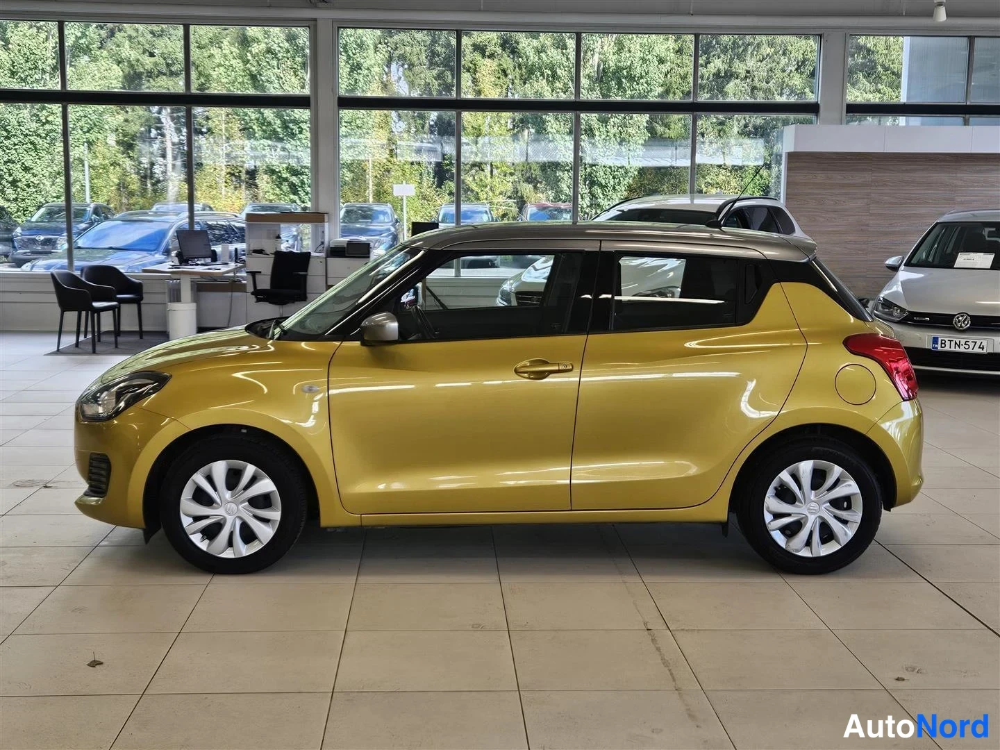 Sivulta: Keltaisen Suzuki Swiftin sivuprofiili vasemmalta nähtynä autosalissa; tumma katto, viisi ovea ja takavalojen reunus näkyvät.