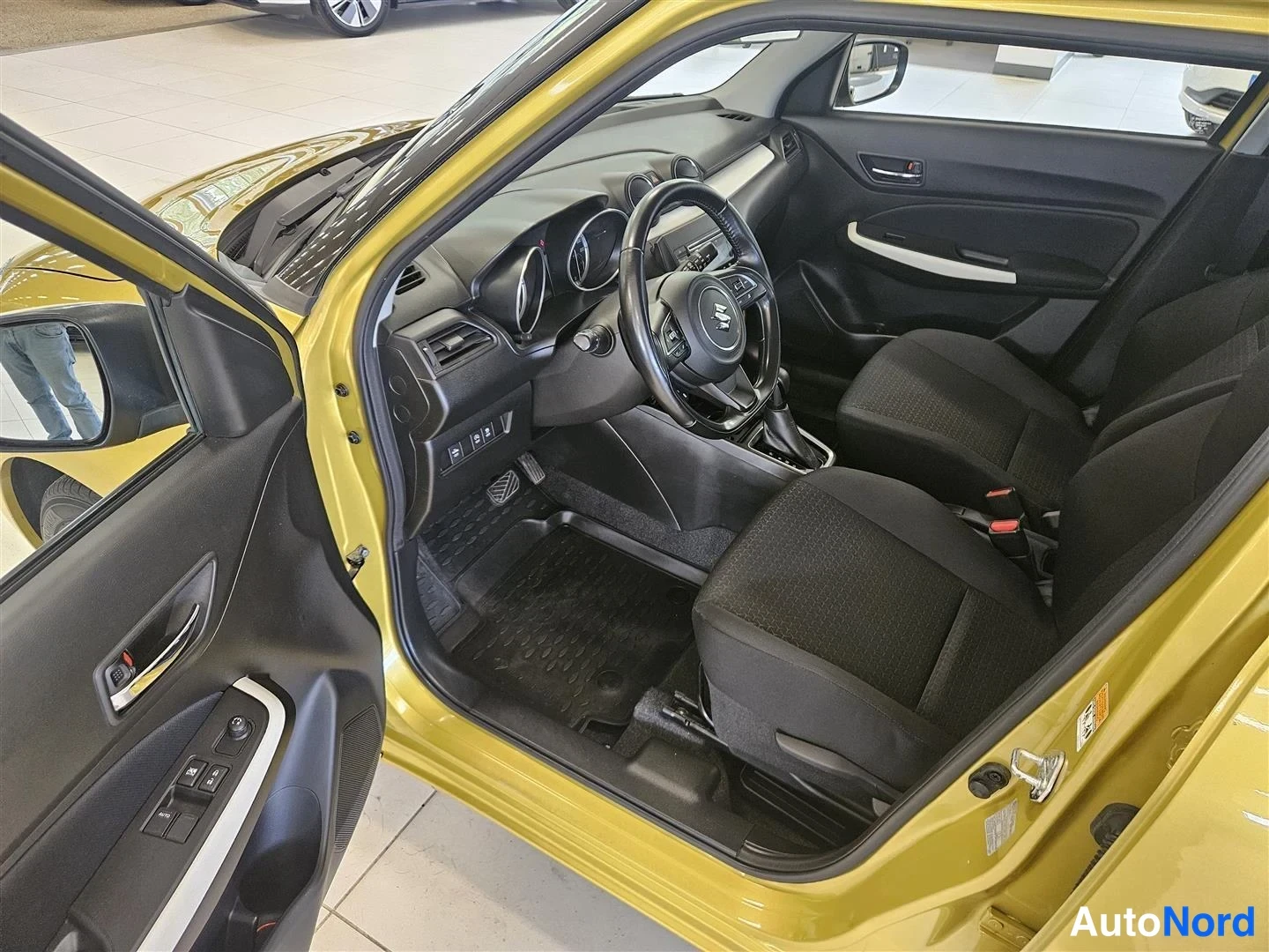 Muu: Suzuki Swiftin sisätilat kuljettajan ovi auki: kojelauta, ohjauspyörä, kangasistuimet ja keltainen koriväri.