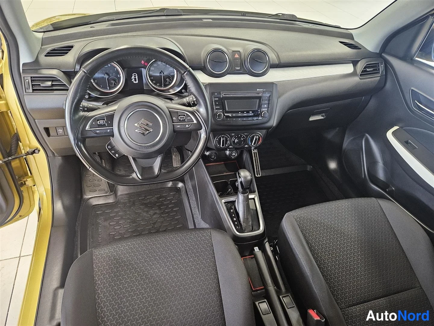 Muu: Suzuki Swiftin ohjaamo edestä: ohjauspyörä, analogiset mittarit, radio ja manuaali-ilmastointi, mustat kangasistuimet.