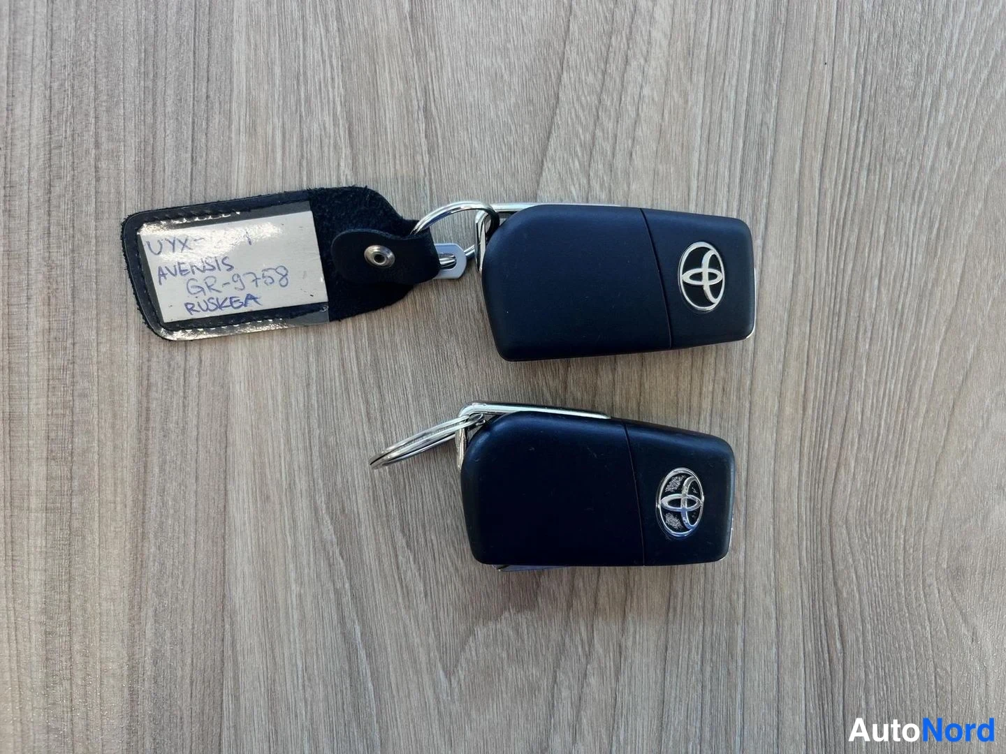Muu: Kaksi Toyota-avainnappia puunkuvioisella pöydällä, mukana käsinkirjoitettu Avensis-kyltti.