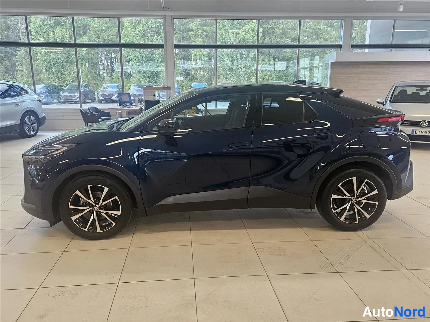 Etuviisto, nokka vasemmalle: Tummansinisen Toyota C-HR:n vasen kylki ja etuosa kolmannespäin sisätiloissa.