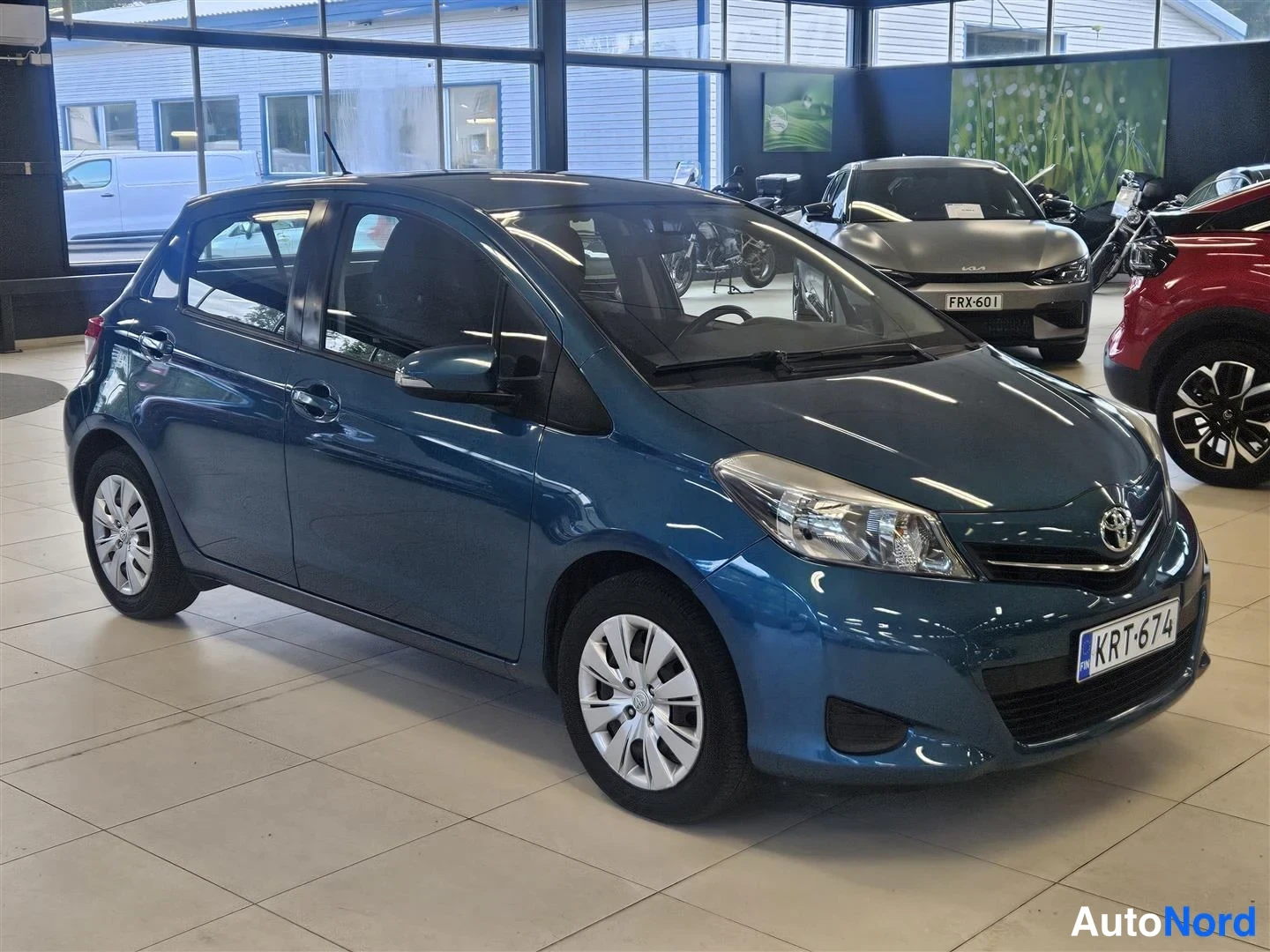 Etuviisto, nokka oikealle: Sinivihreä Toyota Yaris viistosti edestä oikealta myymälätilassa, näkyvissä keula, molemmat kyljet ja rekisteri.
