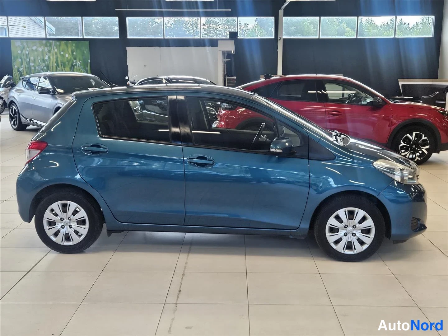 Etuviisto, nokka oikealle: Sinivihreä Toyota Yaris sivusta oikealta, viistosti edestä, näkyvissä kylki, etuvilkku ja keulan osa.