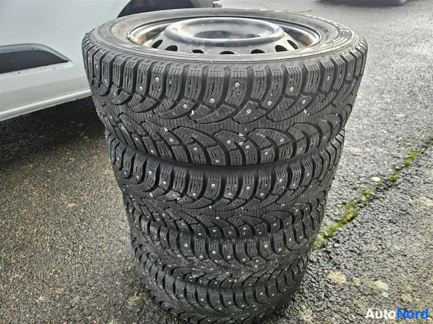 Muu: Pino neljästä nastarenkaasta terävällä metallivanteella; takana valkoisen auton sivua.